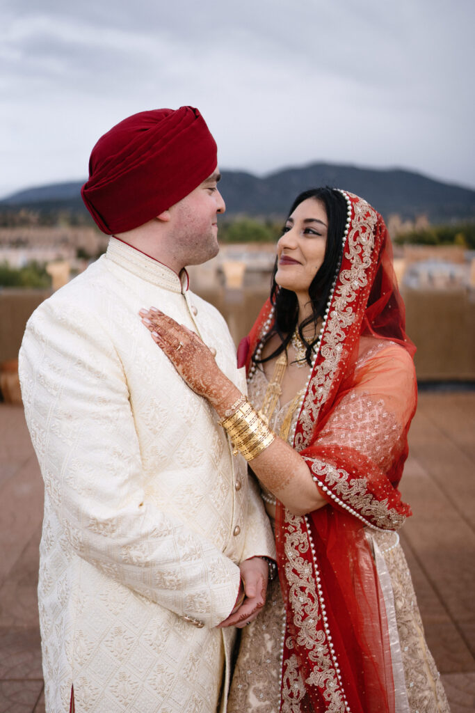 A newlywed couple shares a moment on a rooftop with Santa Fe’s mountains in the background. The groom wears a cream sherwani with a red turban, while the bride’s red dupatta drapes over her shoulder.