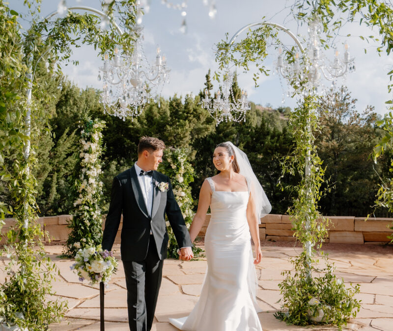 Bride and groom walking down aisle surrounded by lush greenery and chandelier wedding ceremony aisle decor.