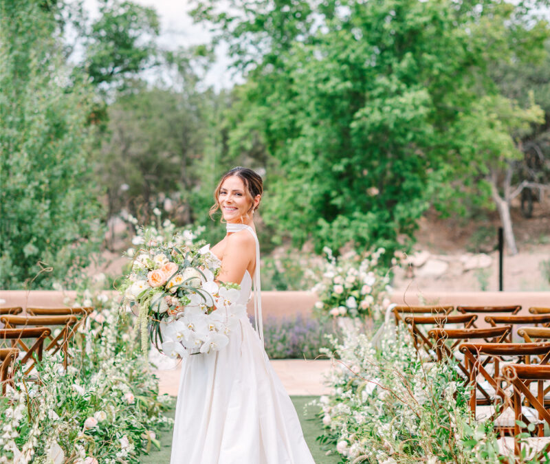 Bride stands in the center of ceremony aisle, holding luxury flower bouquet made for her Santa Fe celebration.